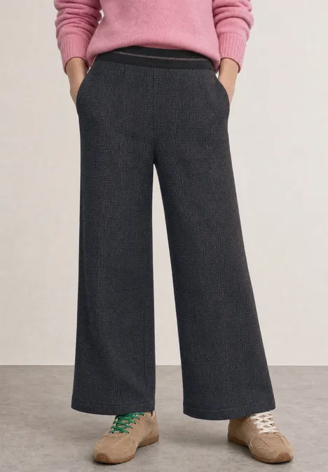 High Waist Wide Leg Hose in Strick-Optik indigo night Person in grauer, weit geschnittener Hose, rosa Pullover, Hände in den Taschen und hellbraune Sneaker.
