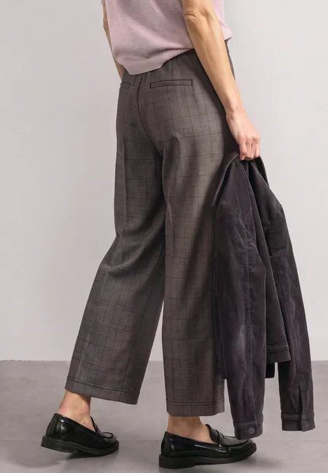 Person trägt weite, karierte Hose, schwarze Loafer und hält eine dunkle Jacke in der Hand.