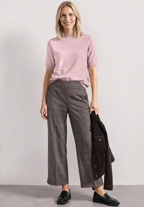 Frau in rosa Pullover und grauen, weiten Hosen hält eine dunkelbraune Jacke in der Hand.
