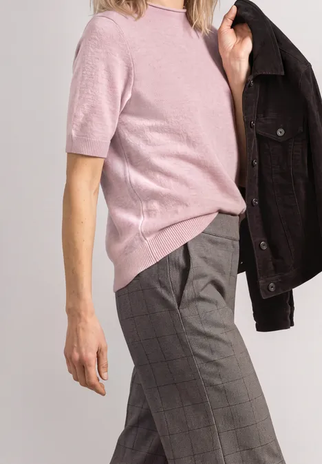 Model in rosa Kurzarm-Pullover, grauen karierten Hosen, schwarze Jacke lässig über die Schulter gelegt.