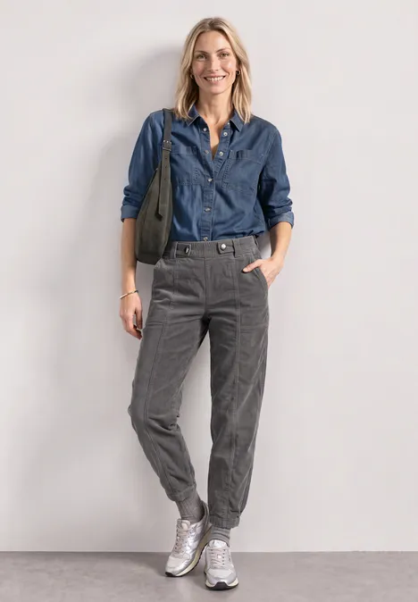 Spodnie High Waist z nogawkami typu barrel z elastycznym pas eclipse grey Uśmiechnięta kobieta w dżinsowej koszuli, szarych spodniach i białych trampkach, z zieloną torbą na ramieniu.