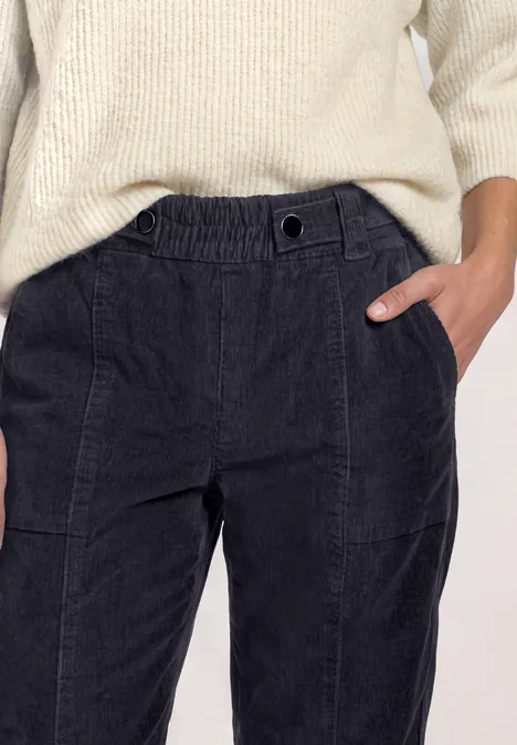 Schwarze Cordhose mit hohem Bund, cremefarbener Strickpullover und Hand in der Hosentasche.