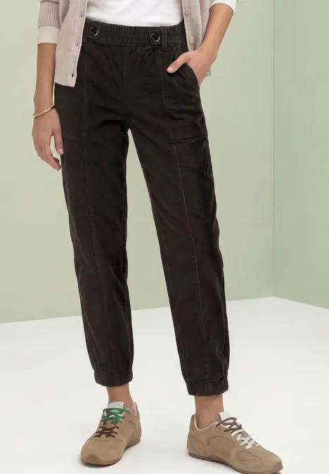 Model draagt zwarte ribfluwelen broek met elastische taille en beige sneakers voor een lichtgroene achtergrond.