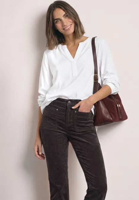Frau in weißer Bluse und brauner Cordhose mit Schultertasche, lächelnd vor hellem Hintergrund.