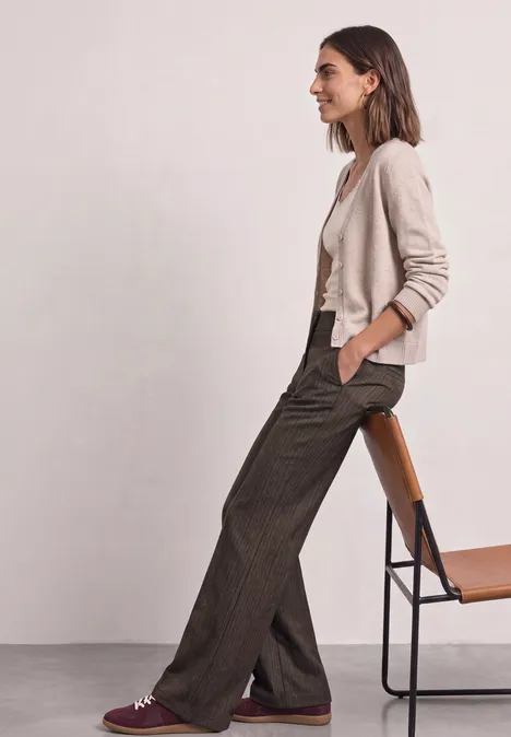 Eine Frau sitzt seitlich auf einem Stuhl, trägt einen hellen Cardigan, weißes Top und weite braune Bundfaltenhose.