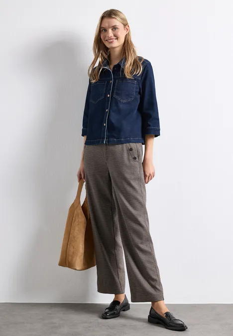 Frau mit dunkelblauer Jeans-Bluse, grauer Hose, schwarzen Loafers und einer braunen Tasche vor weißer Wand.