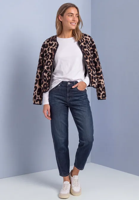 Junge Frau lächelt, trägt Leopardenjacke, weißes Shirt, dunkle Jeans und weiße Sneaker.