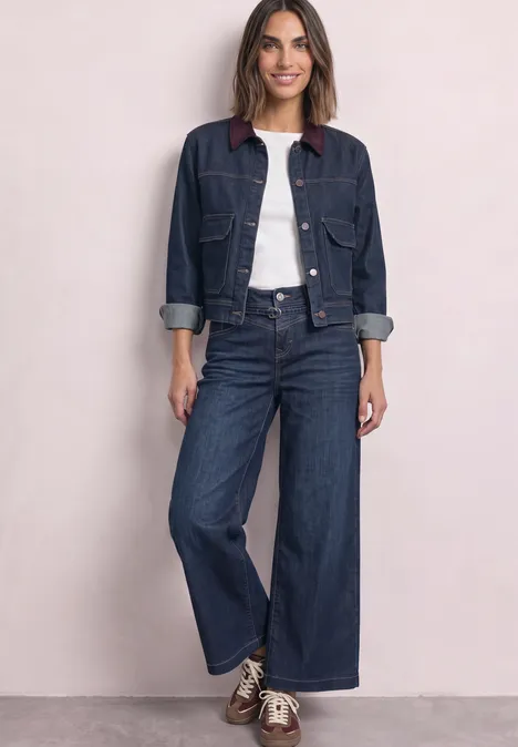 Wide Leg Jeans mit Gürteldetail Dark Blue Washed Frau steht vor hellem Hintergrund in Jeansjacke, weißem Shirt, weiten Jeans und Sneakers.