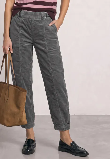 Frau stehend mit grauen Cord-Joggerhosen, rosa Streifenshirt, schwarzen Loafers und brauner Wildledertasche