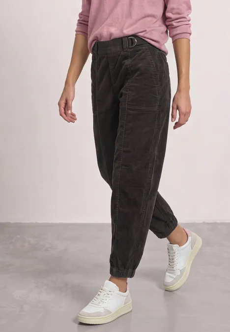 Donkerbruine ribfluwelen broek met losse pasvorm opgerold bij de enkels, gecombineerd met een roze trui en witte sneakers.