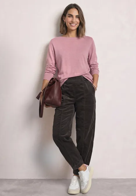 Een vrouw draagt een roze trui, bruine corduroy broek, witte sneakers en een tas.
