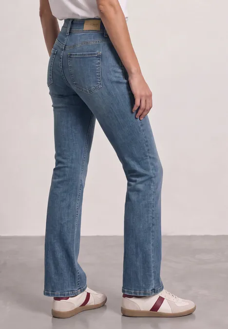High Waist Bootcut Jeans im Slim Fit Mid Blue Washed Frau im geraden, leicht ausgestellten Blue-Jeans, kombiniert mit weißen Loafers, vor schlichtem Hintergrund.