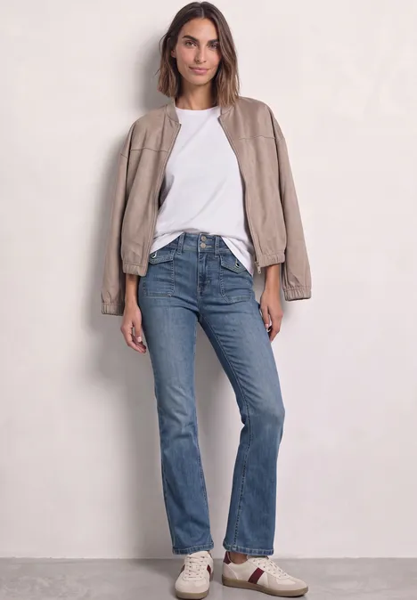 High Waist Bootcut Jeans im Slim Fit Mid Blue Washed Frau trägt beigen Blouson lässig über die Schultern, weißes T-Shirt und blaue Jeans vor neutralem Hintergrund