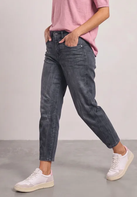 Person in taillierten dunkelgrauen Jeans, weißen Sneakern und rosa Shirt vor neutralem Hintergrund.