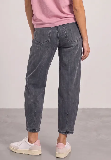 Rückansicht einer Person in lockeren grauen Jeans, rosa Oberteil und weißen Sneakers vor neutralem Hintergrund.