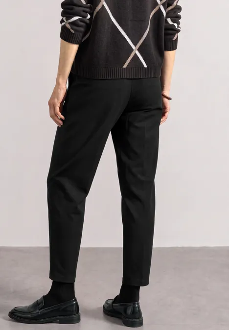 High Waist Barrel Leg Hose im Casual Fit Black Person von hinten in schwarzen Hosen, schwarzen Lederschuhen und dunklem Pullover mit X-Muster.