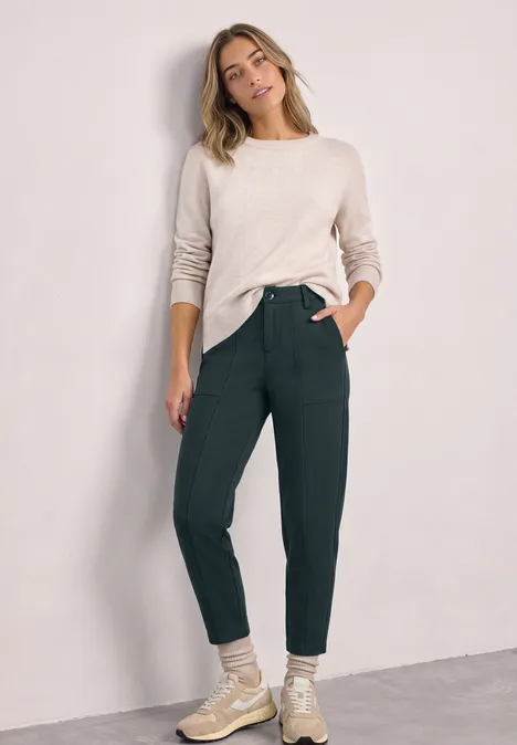 High Waist Barrel Leg Hose im Casual Fit stormy green Eine Frau steht vor einer hellen Wand, trägt beigen Pullover, grüne Hose und helle Schuhe, lächelnd.