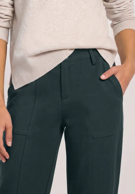 High Waist Barrel Leg Hose im Casual Fit stormy green Weibliche Person in hellem Pullover und dunkler Hose, Hände in den Taschen, Close-up auf Oberkörper und Beine.