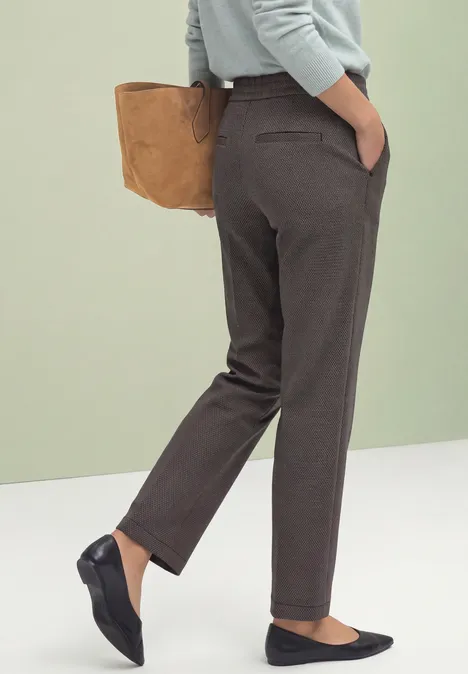 Frau trägt graubraune, gerade geschnittene Hose, schwarze Ballerinas, hält braune Tasche.