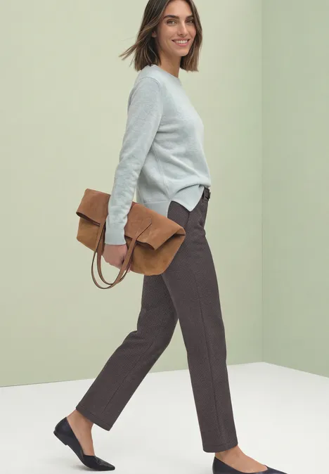 Eine Frau trägt einen hellgrau-blauen Pullover, eine braune Hose und dunkelblaue Slipper, hält eine braune Clutch.