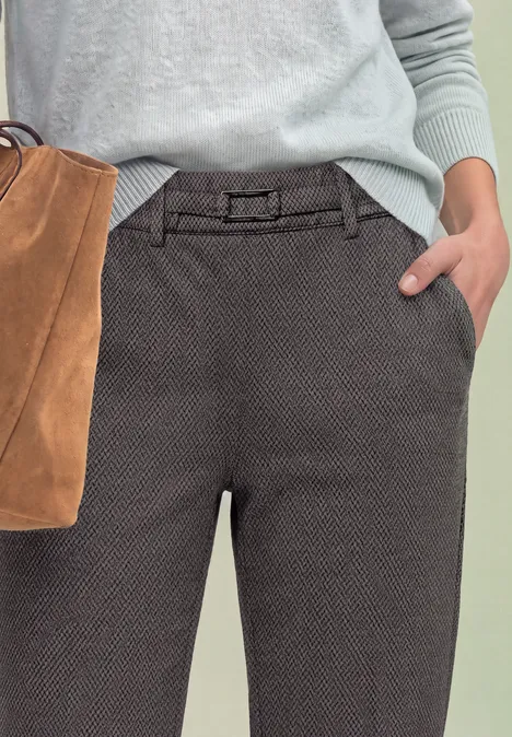 Nahaufnahme: graue, strukturierte Hose mit Gürtel, Hand in Tasche, hellblauer Pullover, braune Wildledertasche.