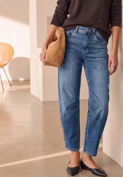 Relaxed Barrel Leg Jeans im Loose Fit Medium Blue Destory Washed Frau trägt gerade geschnittene blaue Jeans, schwarze Ballerinas und beige Clutch vor minimalistischem Interieur.