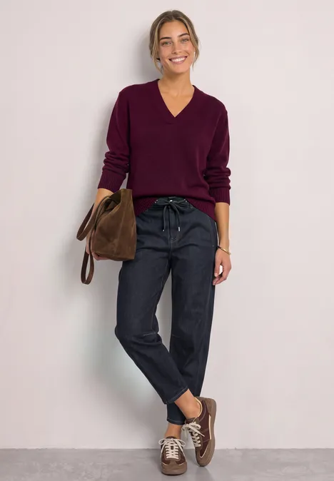 Frau in lila V-Pullover, dunklen Jeans und brauner Tasche steht vor neutralem Hintergrund.