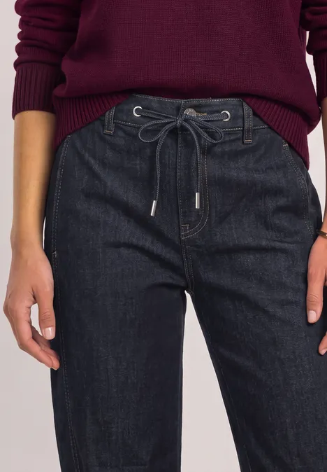 Detailaufnahme einer Frau in dunkelblauen Jeans mit Kordelzug und weinrotem Pullover.