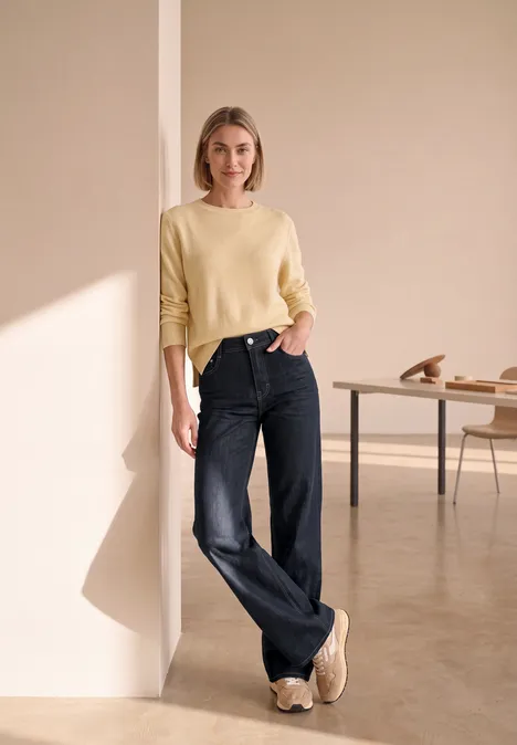 Wide Leg Jeans mit details am Bund Rinse Washed Eine Frau in gelbem Pullover und weiten schwarzen Jeans lehnt sich in einem minimalistischen Raum an die Wand.