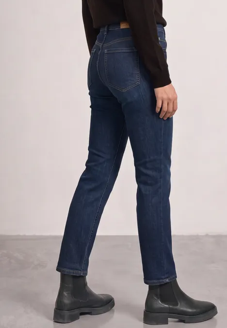 Person trägt blaue Jeans und schwarze Stiefeletten, Aufnahme von der Hüfte abwärts vor neutralem Hintergrund.