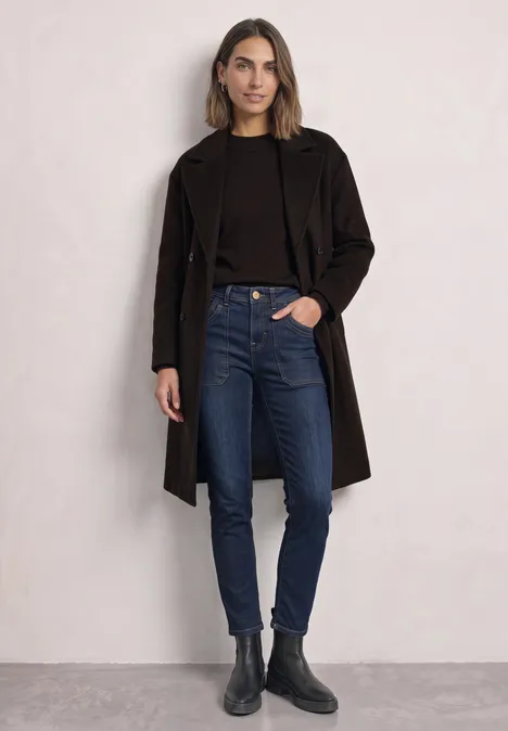 Frau trägt schwarzen Mantel, dunklen Pullover, blaue Jeans und schwarze Stiefeletten vor hellem Hintergrund.