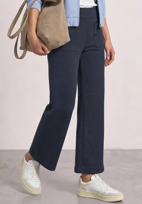 Untersicht einer Person in dunkelblauen, weiten Hosen und weißen Sneakers, mit beiger Tasche.