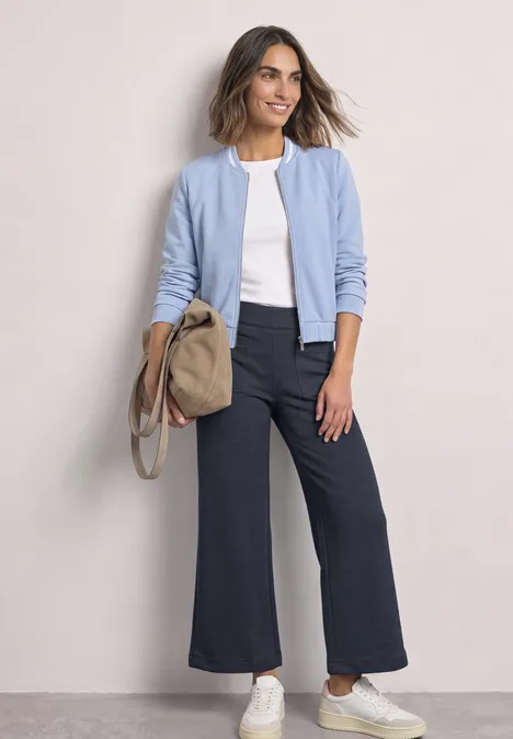 Eine lächelnde Frau trägt eine hellblaue Jacke, weißes T-Shirt, dunkle Schlaghose, weiße Sneakers und eine beige Tasche.
