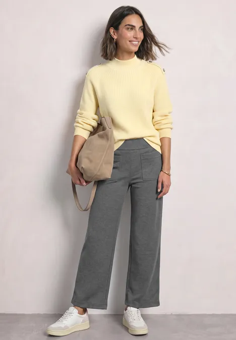 Een vrouw draagt een lichtgele trui, brede grijze broek, witte sneakers en een schoudertas.