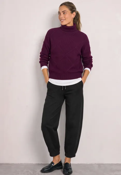 Frau in lila Rollkragenpullover, weißem Hemd und schwarzen, lockeren Hosen mit schwarzen Schuhen.