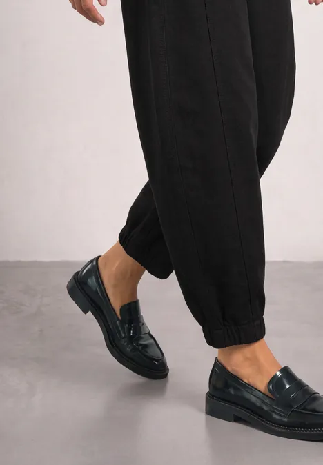 Ein Model trägt schwarze, glänzende Loafers zu hochgeschnittenen Hosen vor neutralem Hintergrund.