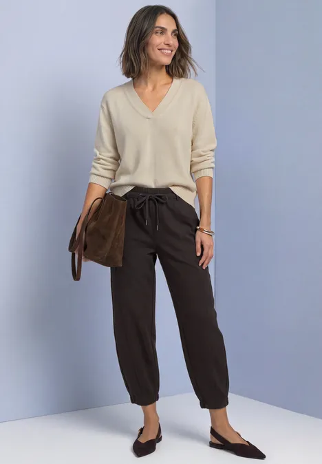Femme en pull beige clair col V, pantalon marron ample et mocassins noirs, tenant pochette marron.