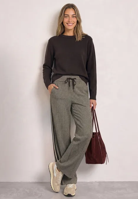 Wide Leg Hose mit Gallonstreifen und Dessin sanded amber Lächelnde Frau in schwarzem Pullover, grauen Jogginghosen und Sneakers hält rote Tasche in lockerer Pose.