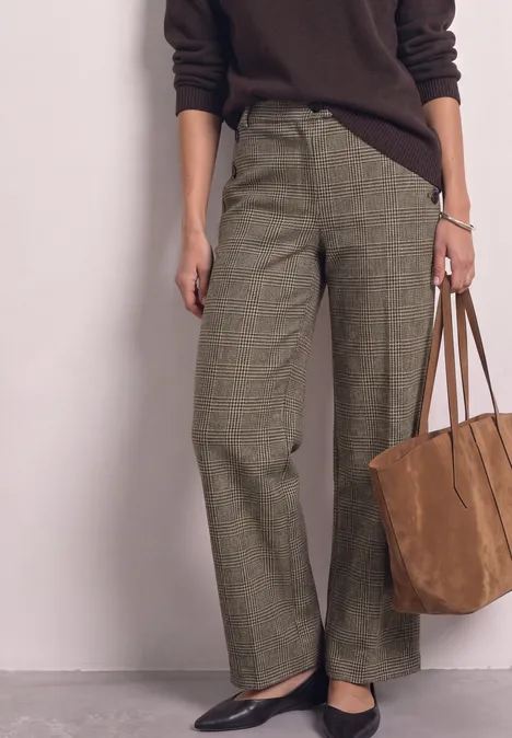 Frau mit braunem Pullover, weiten karrierten Bundfaltenhosen, schwarzen Ballerinas und brauner Wildledertasche.