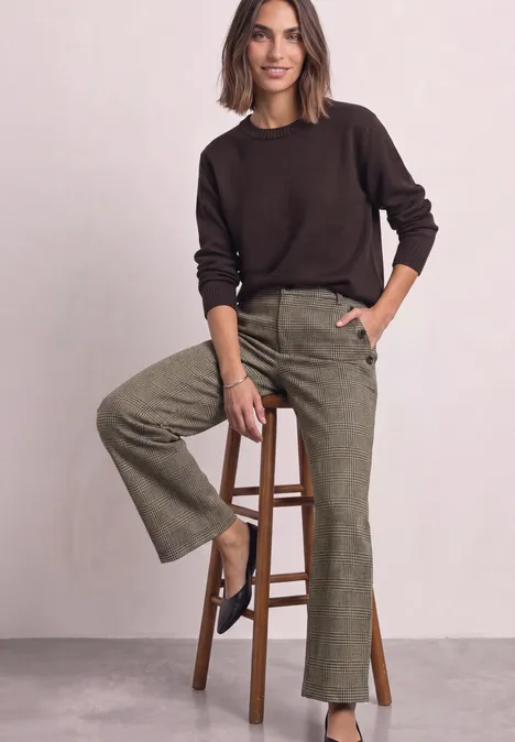 Eine Frau sitzt auf einem Hocker, trägt braunen Pullover und grüne Cordhose vor neutralem Hintergrund.