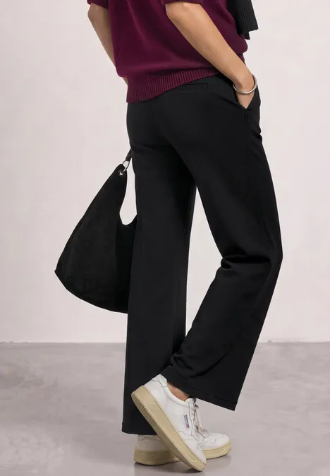 High Waist Wide Leg Hose im Casual Fit Black Seitliche Aufnahme einer Person in schwarzen weiten Hosen, weißen Sneakern, Burgunderpullover und Tasche.