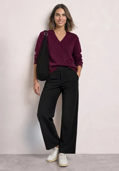 High Waist Wide Leg Hose im Casual Fit Black Frau lehnt lässig an Wand, trägt burgunderfarbenen V-Pullover, schwarze weite Hose, weiße Sneaker und schwarze Tasche.
