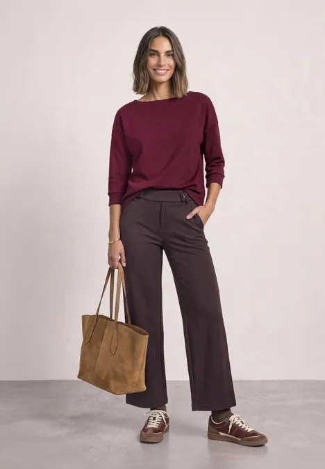 High Waist Wide Leg Hose im Casual Fit ruby vine Frau in burgunderfarbenem Pullover, weitem Hosenrock und Ledertasche vor neutralem Hintergrund.