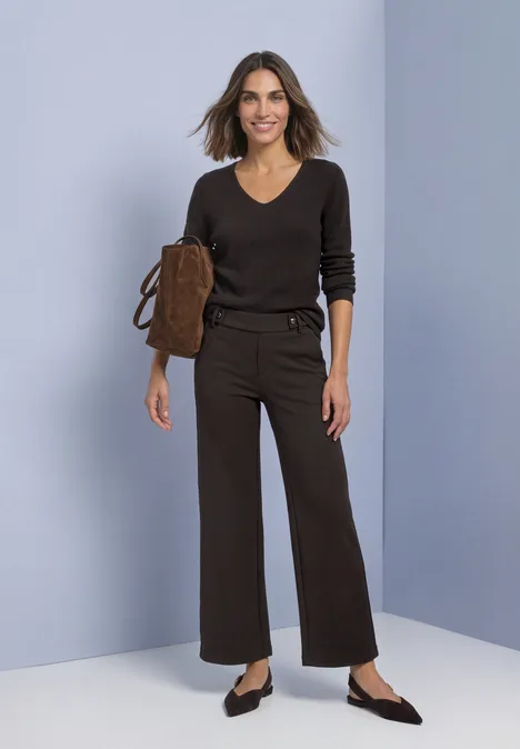 High Waist Wide Leg Hose im Casual Fit black coffee Frau in braunem V-Pullover, weiten Hosen und schwarzen Ballerinas hält eine braune Umhängetasche vor hellem Hintergrund.