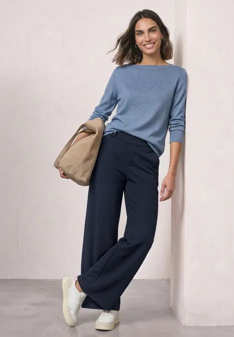 High Waist Wide Leg Hose im Casual Fit darkest blue Frau in blauem Pullover und weitem dunkelblauen Hosen mit weißen Sneakern und Beuteltasche, lässig an Wand gelehnt.