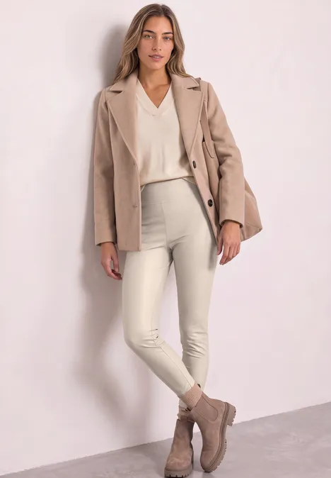 Vrouw in beige blazer, ivoorkleurige trui, witte broek en taupe enkellaarsjes tegen een lichte muur.