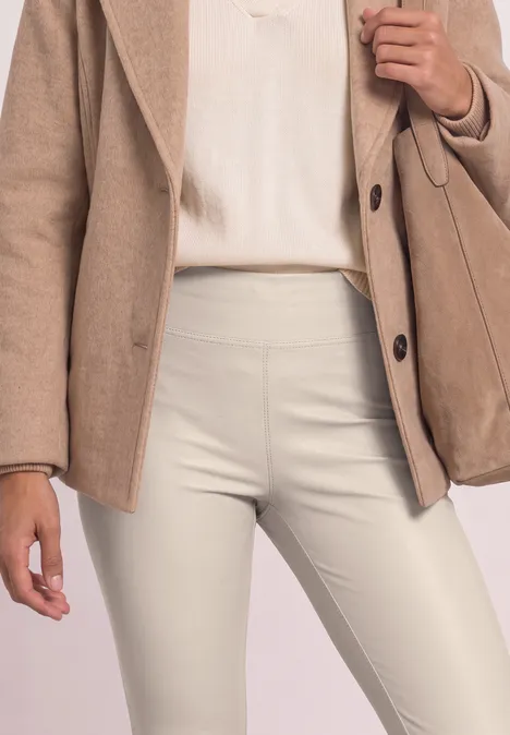 Close-up van een persoon met een beige blazer, licht topje en crèmekleurige, nauwsluitende broek met bijpassende tas.