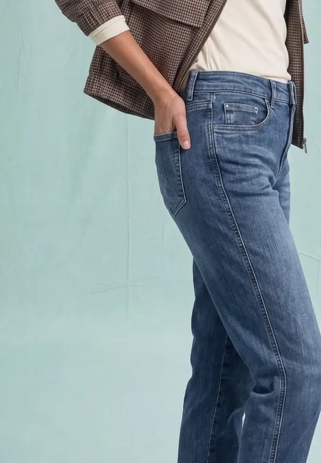 Eine Person trägt blaue Jeans und eine braune Jacke, steht seitlich mit Hand in Tasche.