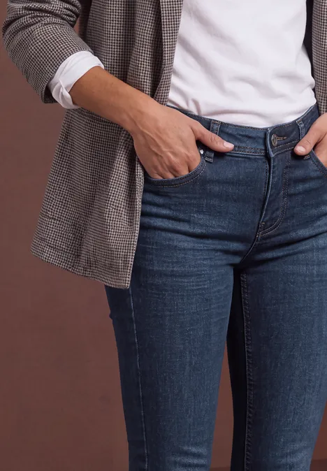 Eine Person in weißem Shirt, kariertem Blazer und blauen Jeans mit Händen in den Taschen.