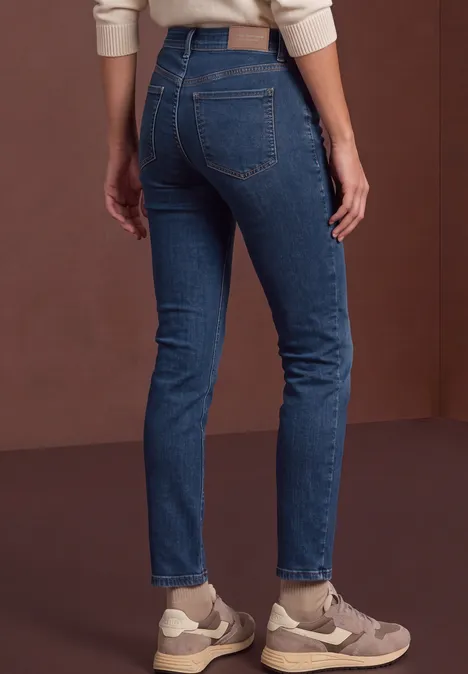 Mid Waist Straight Leg Jeans w kroju Slim Fit dark blue soft wash Ujęcie z tyłu przedstawiające osobę w niebieskich dżinsach i trampkach na brązowym tle.